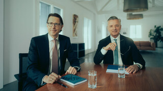 Zwei Personen in Business-Kleidung sitzen an einem Besprechungstisch und blicken an der Kamera vorbei.