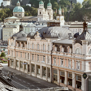 Stammhaus des Bankhaus Spängler in der Schwarzstraße 1 in 5020 Salzburg von außen. Die Fassade ist hellrosa, auffällig ist das Kuppeldach. Im Hintergrund sieht man die Festung Hohensalzburg sowie die Kuppeln des Salzburger Doms. 