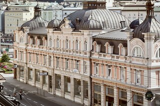 Stammhaus des Bankhaus Spängler in der Schwarzstraße 1 in 5020 Salzburg von außen. Die Fassade ist hellrosa, auffällig ist das Kuppeldach. Im Hintergrund sieht man die Festung Hohensalzburg sowie die Kuppeln des Salzburger Doms. 