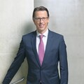 Ein lächelnder Herr in Geschäftskleidung steht vor einer Betonwand.