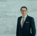 Ein lächelnder Herr in Geschäftskleidung steht vor einer Betonwand.