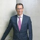 Ein lächelnder Herr in Geschäftskleidung steht vor einer Betonwand.