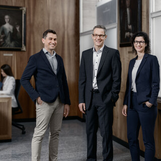 Eine Dame und zwei Herren in Geschäftskleidung stehen lächelnd in einem Büro.