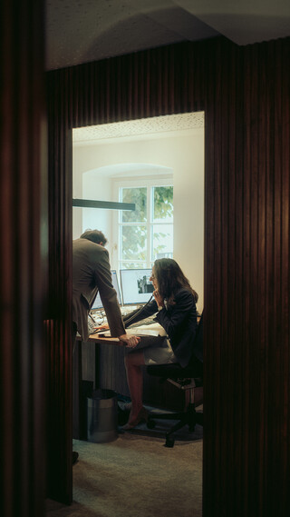 Zwei Personen befinden sich gemeinsam im Büro: Ein Mann lehnt am Tisch, während die Frau telefoniert. Die Aufnahme entstand aus der Ferne, durch die Tür hindurch.