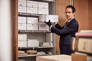 Junger Mann in Anzug steht vor einer Schrankwand und nimmt einen Karton mit Unterlagen heraus.