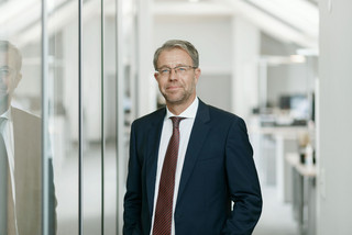 Mann mit leichtem Lächeln steht in einem Bürogang. Er hat schwarz-grau melierte Haare und trägt eine eckige Brille. Gekleidet ist er in ein schwarzes Sakko mit weißem Hemd und grau-rot-gestreifter Krawatte.