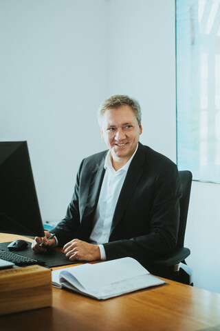 Ein lächelnder Herr in Geschäftskleidung sitzt an einem modernen Schreibtisch.