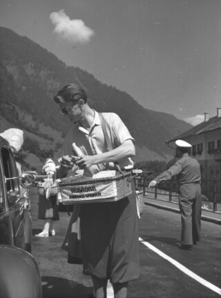 Ein historisches Schwarz-Weiß-Foto zeigt einen sommerlich bekleideten Herrn mit einem umgehängten Korb mit dem er auf einer Straße Geldwechsel anbietet.