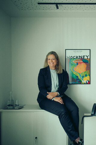 Eine blonde Frau lächelt. Sie trägt eine weiße Bluse mit Rüschen und einen dunklen Blazer und sitzt auf einem Sideboard.