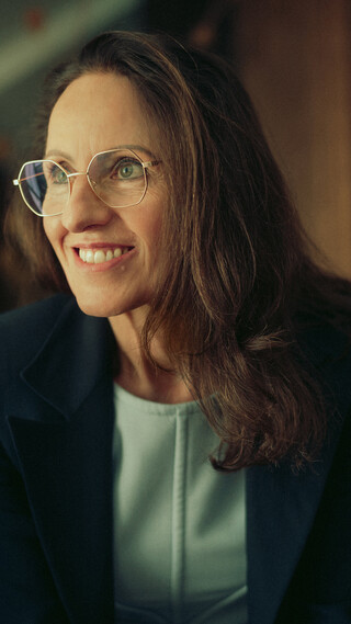 Eine Frau mittleren Alters mit dunklen schulterlangen Haaren, hellblauem Oberteil, dunklem Blazer und Brille lächelt und blickt nach rechts; Aufnahme zeigt den Oberkörper.