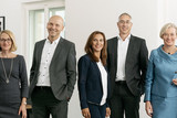 5 Personen (3 Frauen, 2 Männer) in schicker Kleidung befinden sich in einem Büro und lächeln freundlich.