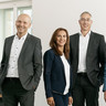 5 Personen (3 Frauen, 2 Männer) in schicker Kleidung befinden sich in einem Büro und lächeln freundlich.