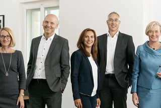 5 Personen (3 Frauen, 2 Männer) in schicker Kleidung befinden sich in einem Büro und lächeln freundlich.