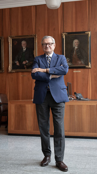 Mann mit Brille, Anzug und verschränkten Händen steht in einem Büro mit dunkler Holzverkleidung. Im Hintergrund sind 4 Gemälde/ Portraits erkennbar.