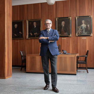 Mann mit Brille, Anzug und verschränkten Händen steht in einem Büro mit dunkler Holzverkleidung. Im Hintergrund sind 4 Gemälde/ Portraits erkennbar.