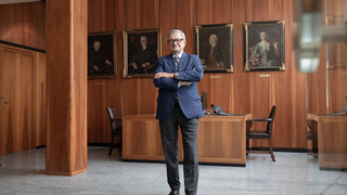 Mann mit Brille, Anzug und verschränkten Händen steht in einem Büro mit dunkler Holzverkleidung. Im Hintergrund sind 4 Gemälde/ Portraits erkennbar.