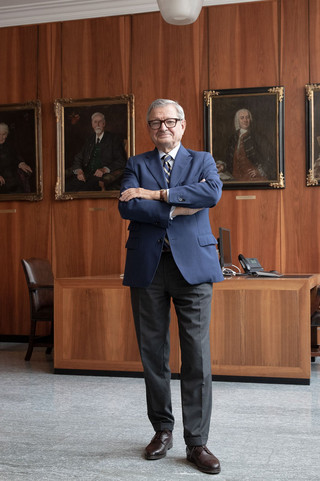 Mann mit Brille, Anzug und verschränkten Händen steht in einem Büro mit dunkler Holzverkleidung. Im Hintergrund sind 4 Gemälde/ Portraits erkennbar.