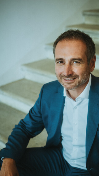 Ein lächelnder Herr in Anzug sitzt auf einer historischen Treppe.