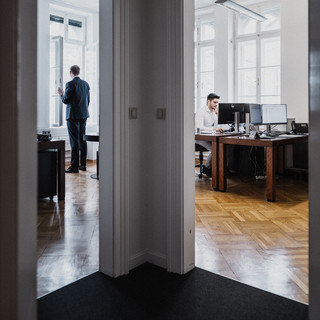 Blick in zwei Büros vom Gang aus. Im linken Büro steht ein Mann am Fenster. Im rechten Büro sitzt ein Mann am Computer.