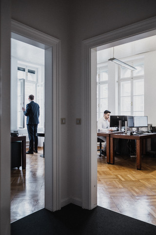 Blick in zwei Büros vom Gang aus. Im linken Büro steht ein Mann am Fenster. Im rechten Büro sitzt ein Mann am Computer.