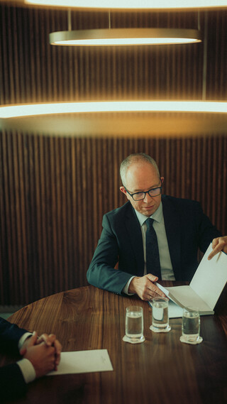 Ein Mann sitzt im Besprechungsraum und blättert in einer Broschüre; von einer zweiten Person sind nur die Hände sichtbar.