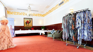 Großer Raum mit rottem Teppichboden mit bunten Kleidern auf einer Kleiderstange, zwei grünen Stühlen und einer Schaufensterpuppe mit einem rosa-farbenen Federnkleid sind ersichtlich.