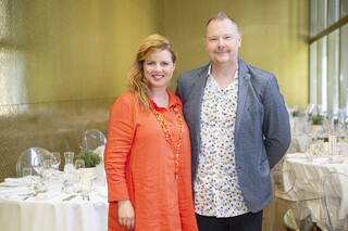 Frau in orangenem Kleid mit blonden langen Haaren und Herr in buntem Hemd und blauem Sakko stehen vor festlich gedeckten Tischen.