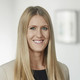 Lächelnde Frau mit blonden, langen Haaren und blauen Augen trägt ein schwarzes Oberteil und einen beige-farbigen Blazer. 