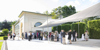 Weiße Orangerie von außen, Gäste vor dem Gebäude. Es herrscht schönes Wetter.