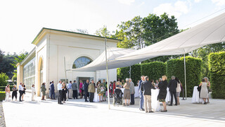 Weiße Orangerie von außen, Gäste vor dem Gebäude. Es herrscht schönes Wetter.