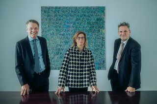 Drei Personen stehen an einem dunklen Besprechungstisch und blicken in die Kamera.